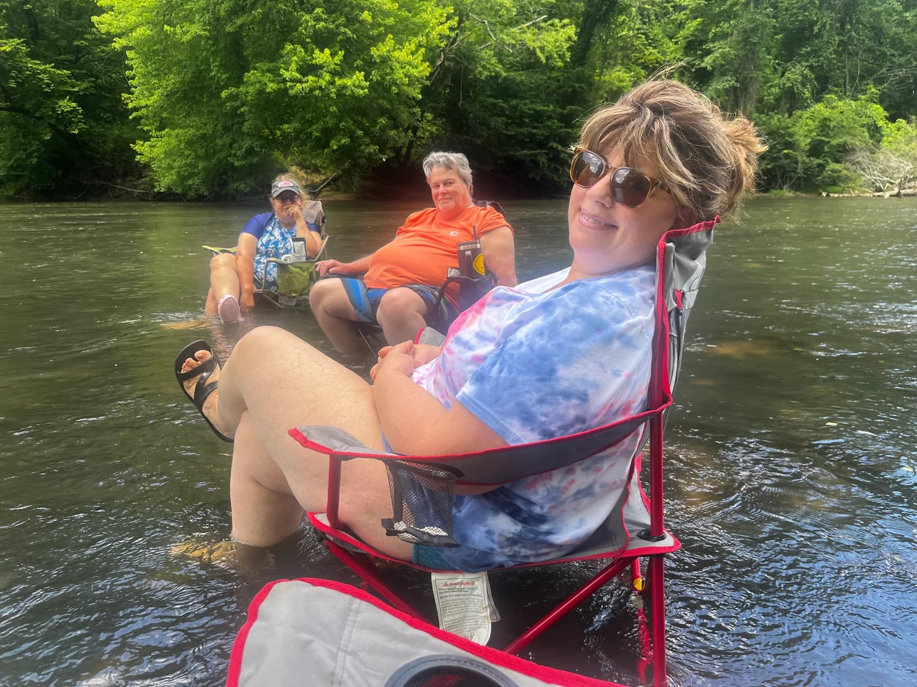 Three adults relaxing in folding chairs in the river beneath leafy trees